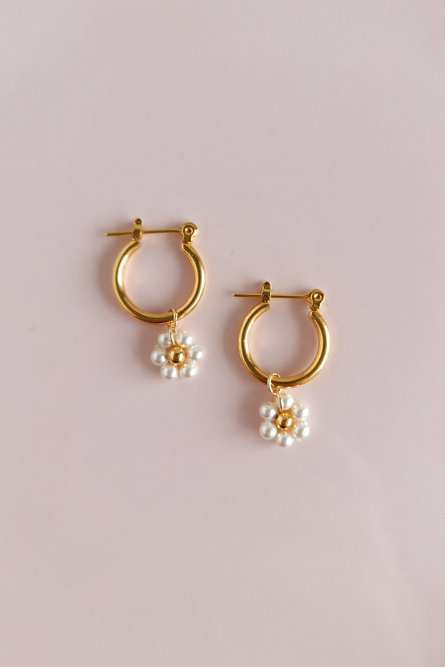 A close-up photo of a pair of gold hoop earrings. The earrings feature delicate pearl clusters resembling daisies with tiny gold bead centers. The background is pink.