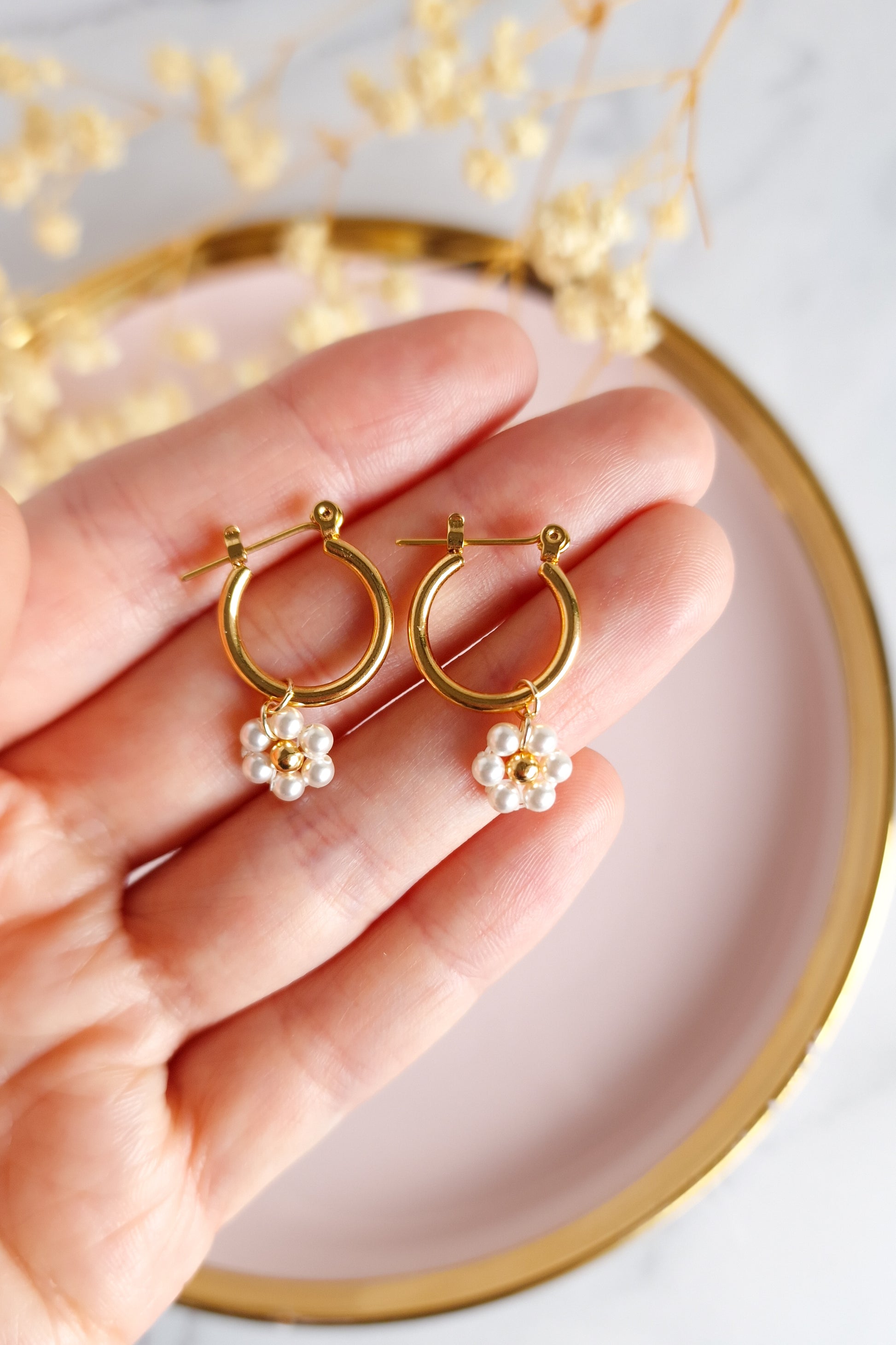 A close-up photo of a hand holding a pair of gold hoop earrings. The earrings feature delicate pearl clusters resembling small flowers with tiny gold bead centers.