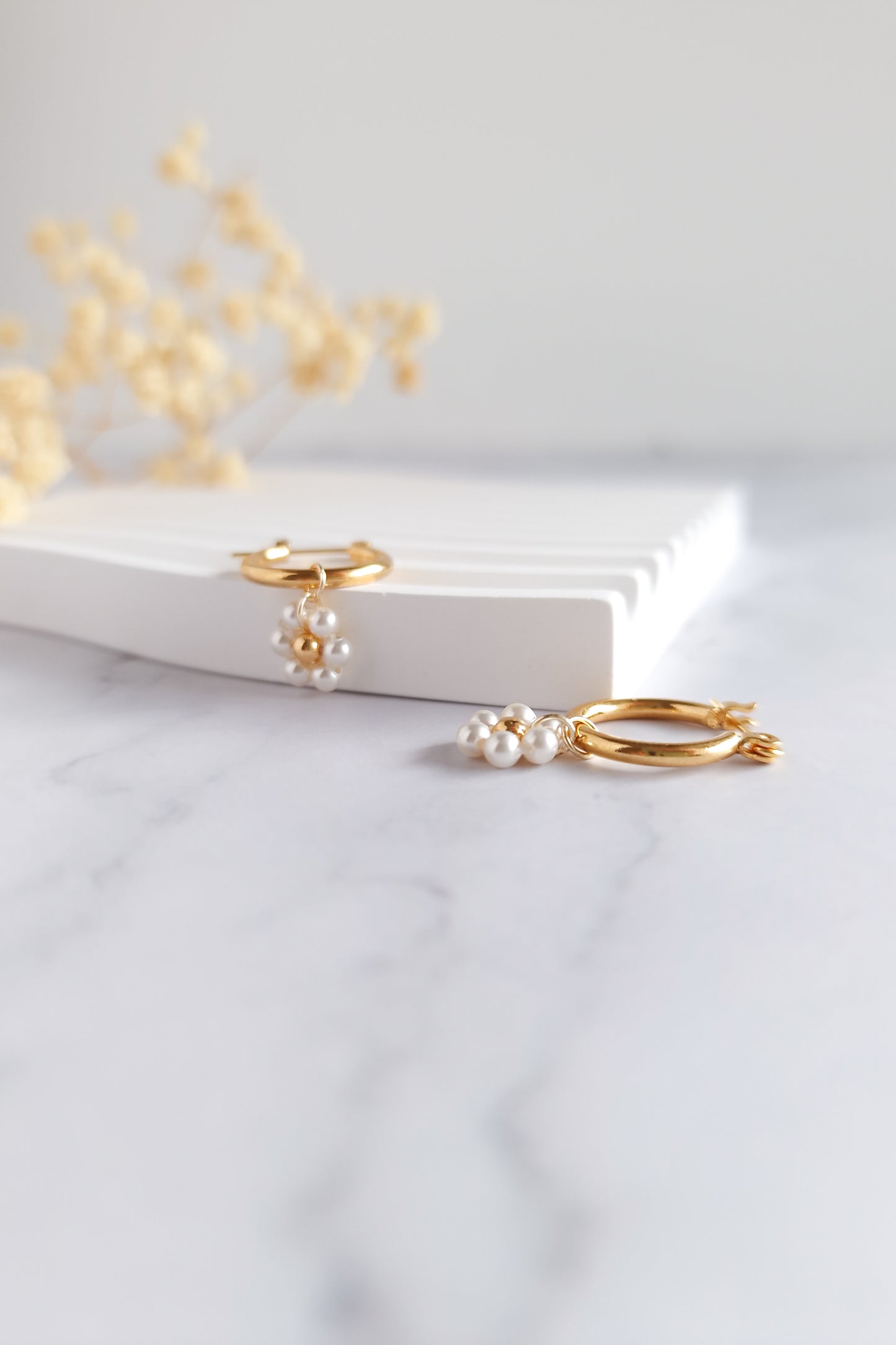 A close-up of a pair of gold hoop earrings with delicate pearl clusters that resemble small flowers. The centers of the flowers are tiny gold beads.