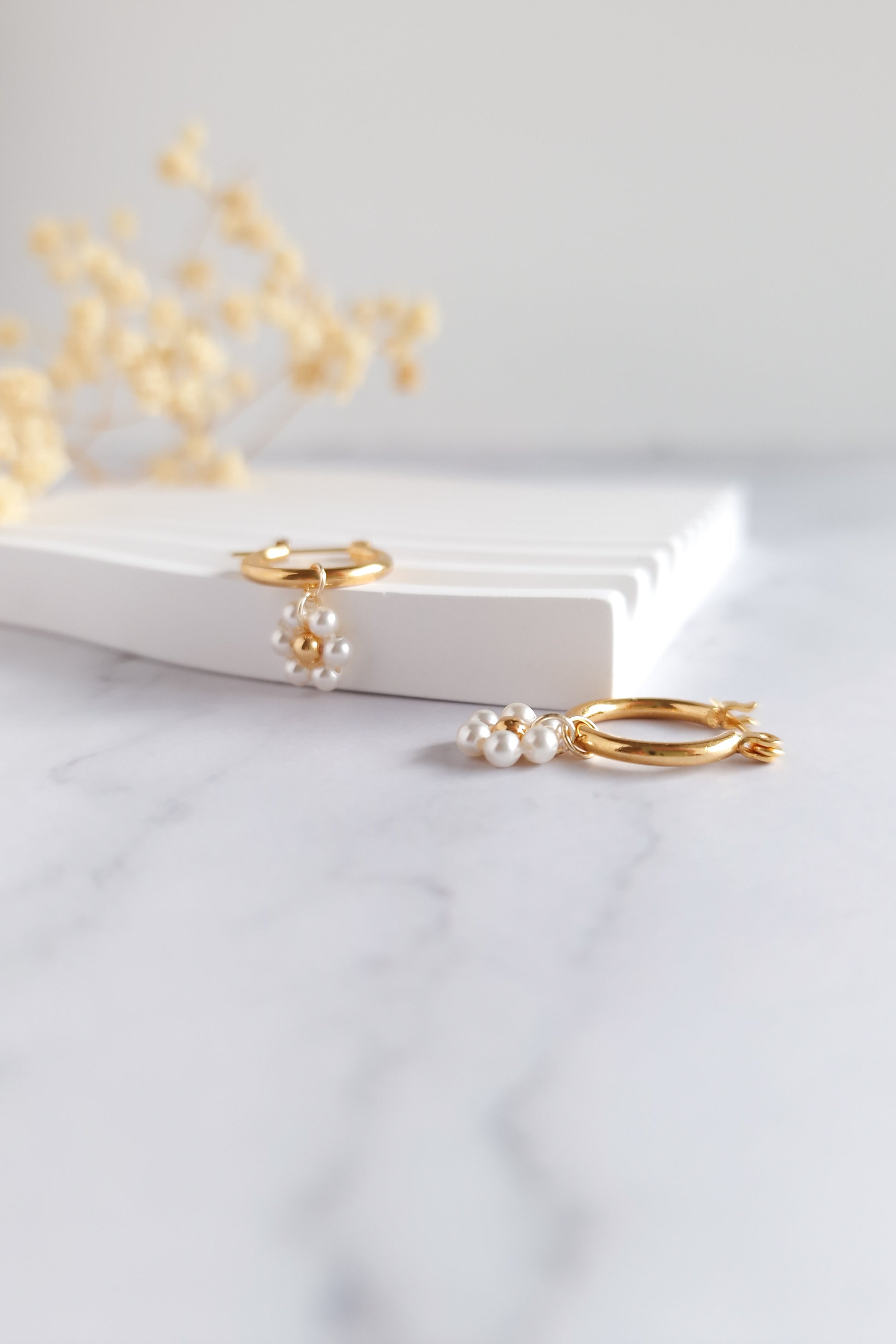 A close-up of a pair of gold hoop earrings with delicate pearl clusters that resemble small flowers. The centers of the flowers are tiny gold beads.